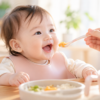 明るいダイニングで、スプーンから離乳食を食べようと笑顔で口を開ける生後8か月ごろの赤ちゃん