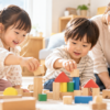 積み木で一緒に遊ぶ3歳ごろの女の子と男の子を、そばで優しく見守る大人