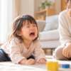 リビングの床で泣いて癇癪を起こす3歳ごろの子どもと、そばで穏やかに見守る母親