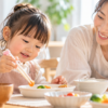 初めてのお箸で食べ物をつかもうと頑張る3歳ごろの女の子と、優しく見守る母親