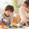 リビングで積み木遊びに夢中になる3歳ごろの男の子と、そばで優しく声をかける母親